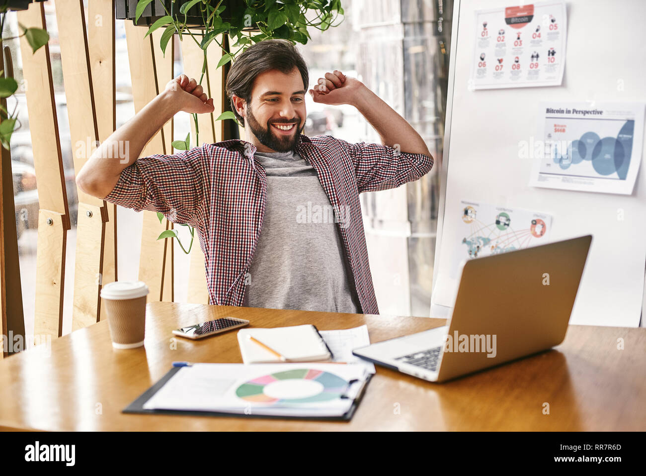 Young businessman in casual wear stretching his arms and body for relaxing while working in office with laptop. A cup of coffee, smartphone, notebook on wooden table. Resting for prevent office syndrome Stock Photo