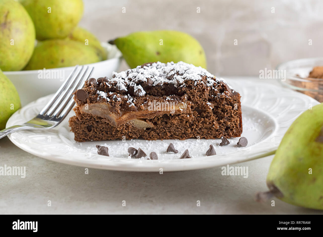 Close up side view of Chocolate Pear Cake sprinkled with powdered sugar on white plate.  Chocolate chips and fork on plate.  Pears and cocoa powder in Stock Photo