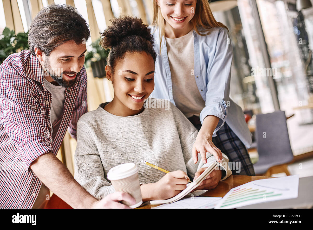 Young beautiful african american woman sitting over desk and making notes while taking advice from her colleagues. They are standing beside her and help to think of new ideas Stock Photo