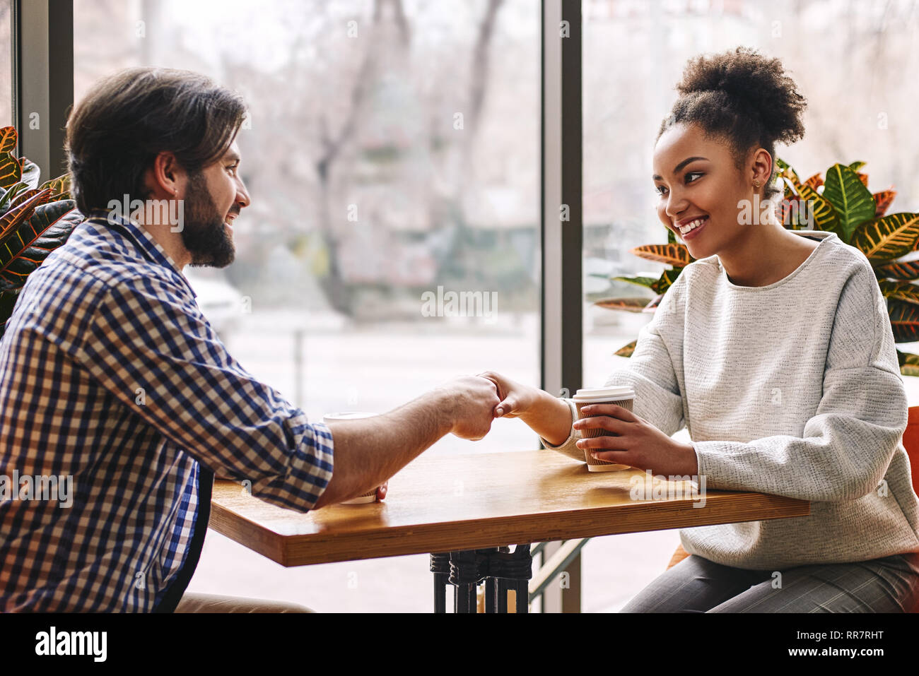 Side view portrait of two successful business people, man and woman, smiling and shaking hands across table during meeting in cafe. African american woman and caucasian dark-haired man Stock Photo