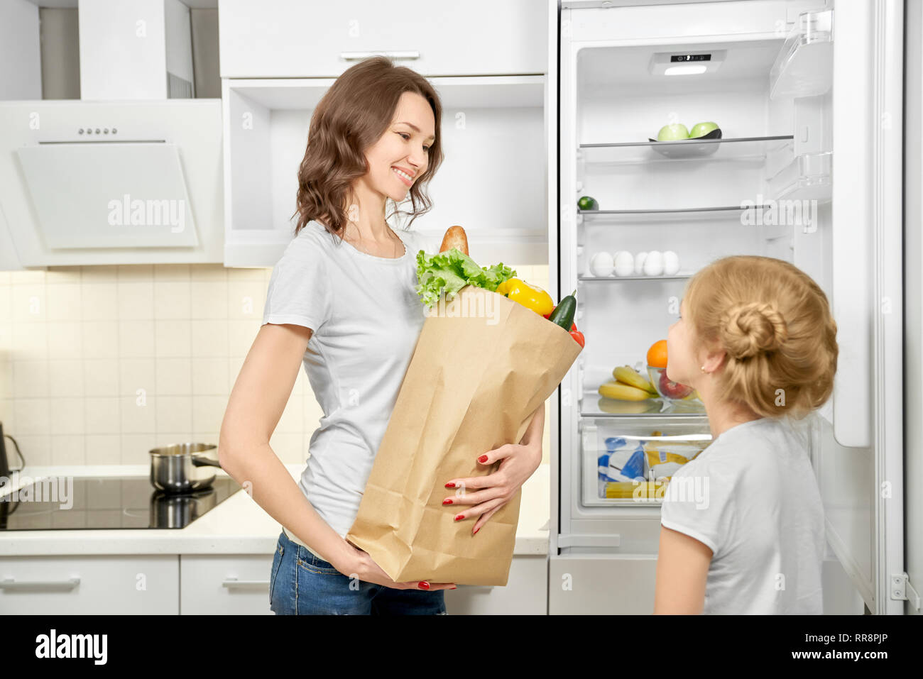 Mother standing in modern kitchen, holding big paper bag with vegetables, greens and baguette. Daughter standing near empty fridge. Fresh products aft Stock Photo