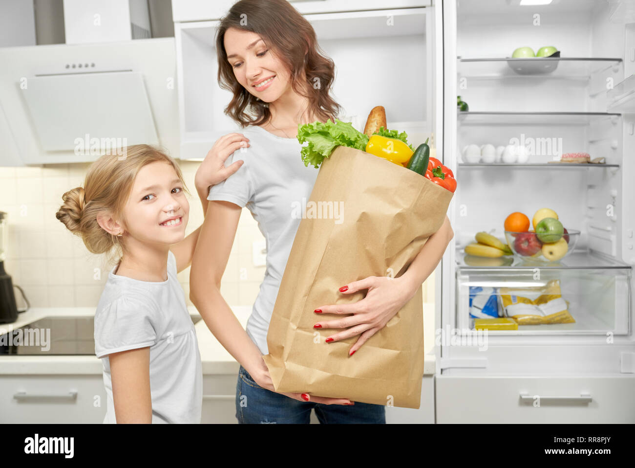 Happy, cheerful mother and daughter standing and posing in kitchen near empty fridge. Mom holding big paper bag full of fresh products after shopping, Stock Photo