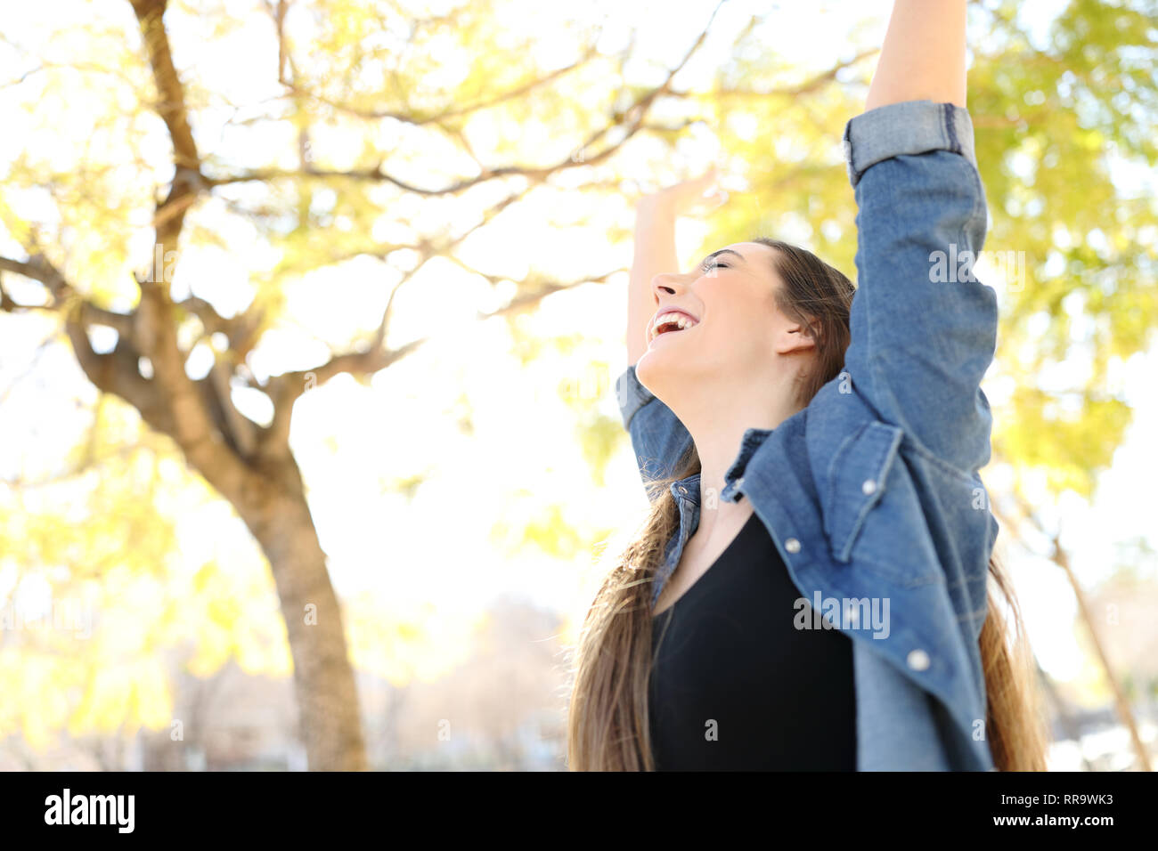 Excited woman raising arms celebrating success in a park with trees in ...