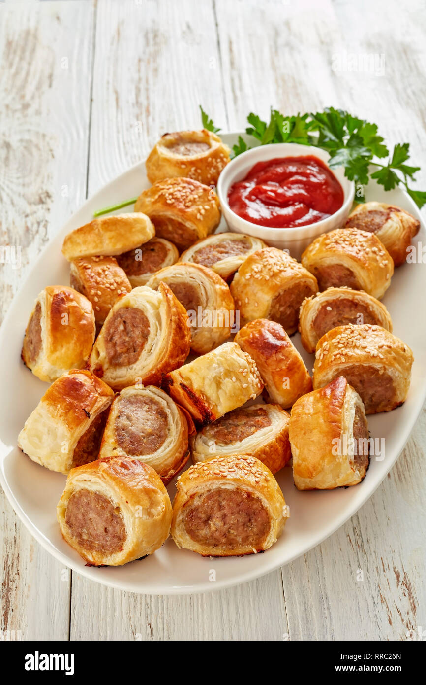 freshly baked Puff pastry Sausage rolls with ketchup and parsley on a white plate, english party food, vertical view from above, close-up Stock Photo