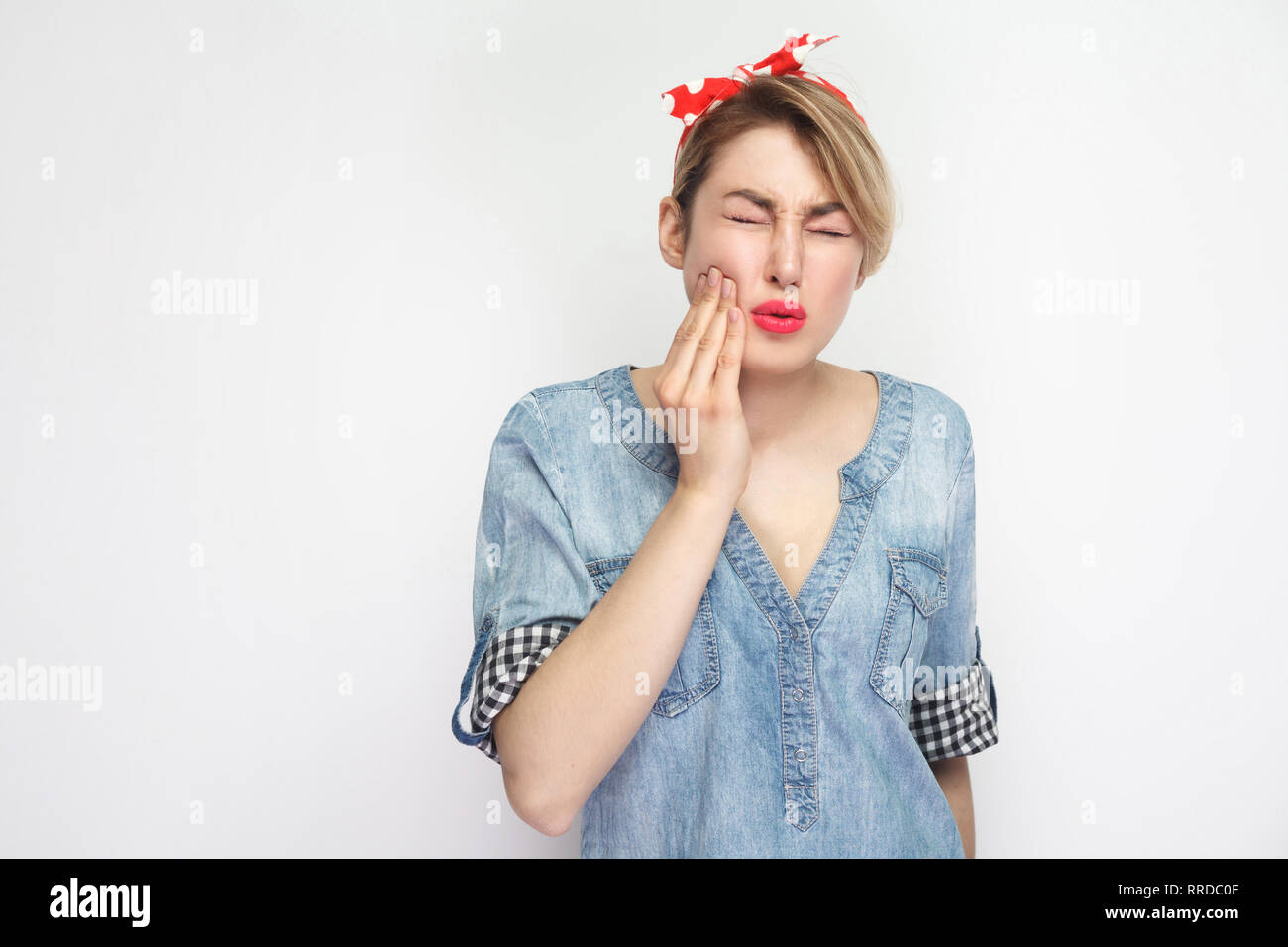 Tooth ache or pain. Portrait of young woman in casual blue denim shirt with makeup and red headband standing and feeling pain on her tooth. indoor stu Stock Photo
