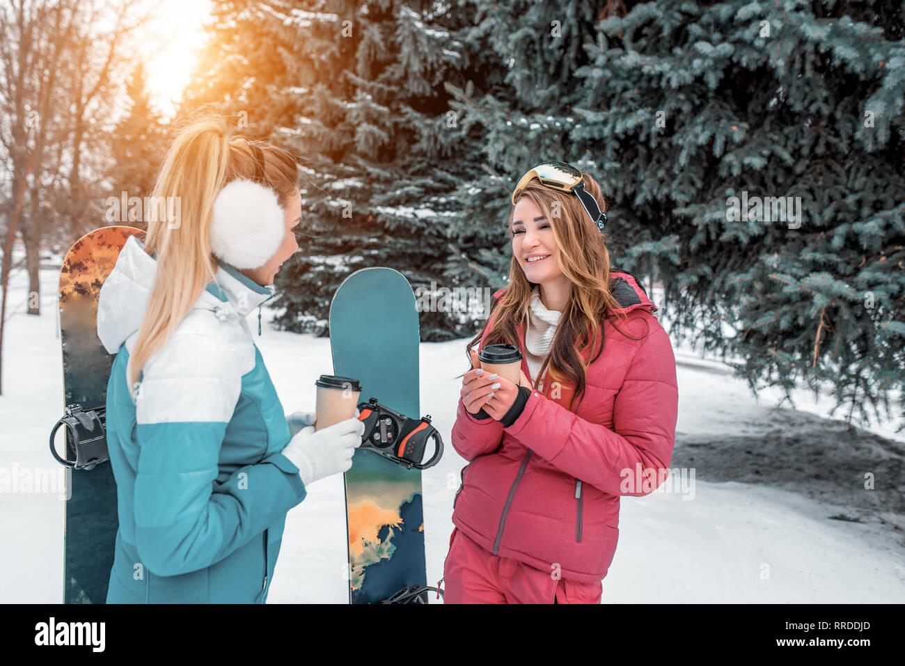 Two girls girlfriends on background of snowboards, talking winter park background of green Christmas trees. In hands of coffee mugs tea. Concept best Stock Photo