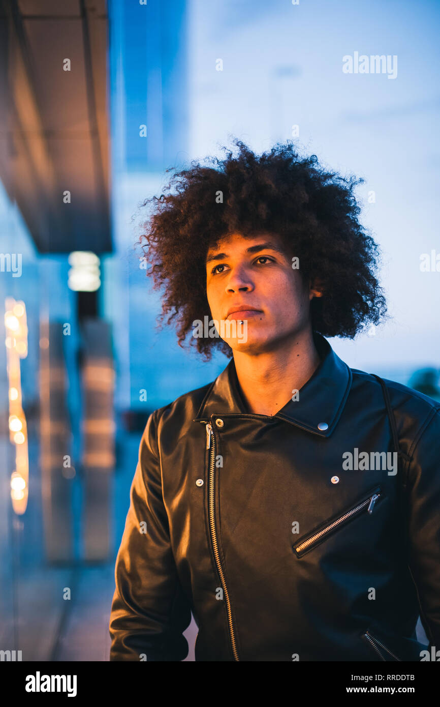 Handsome young man with afro hair illuminated by yellow light of a showcase looking seriously with urban background in blue tones Stock Photo