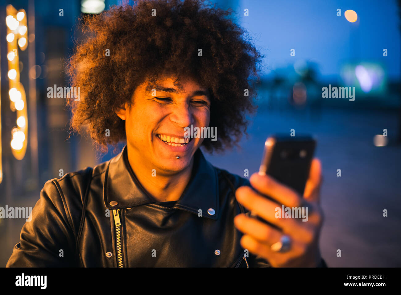 Stylish young man with afro hair smiling by using smart-phone in the street at night, illuminated by the lights of a showcase Stock Photo