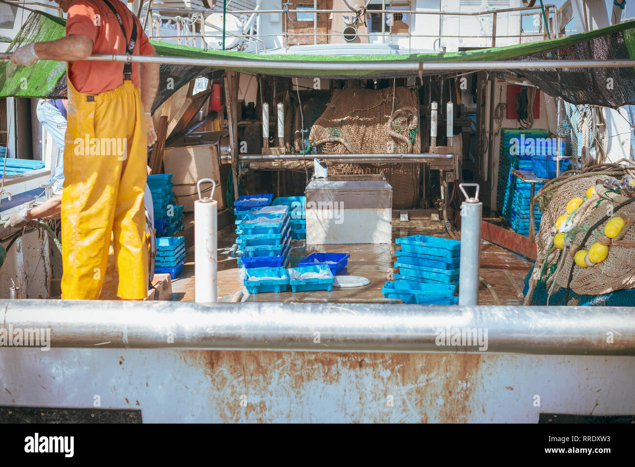 A fisherman wearing yellow overalls tidies up equipment on the deck of a fishing trawler at the port in Denia, Spain. Stock Photo