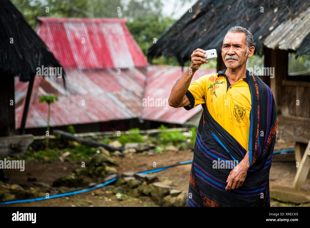 A man takes a photo with his smartphone, his traditional ikat sarong draped over a yellow polo shirt, houses with rust-red corrugated metal roofs behind him in a mountain village near Moni, Flores, Indonesia. Stock Photo