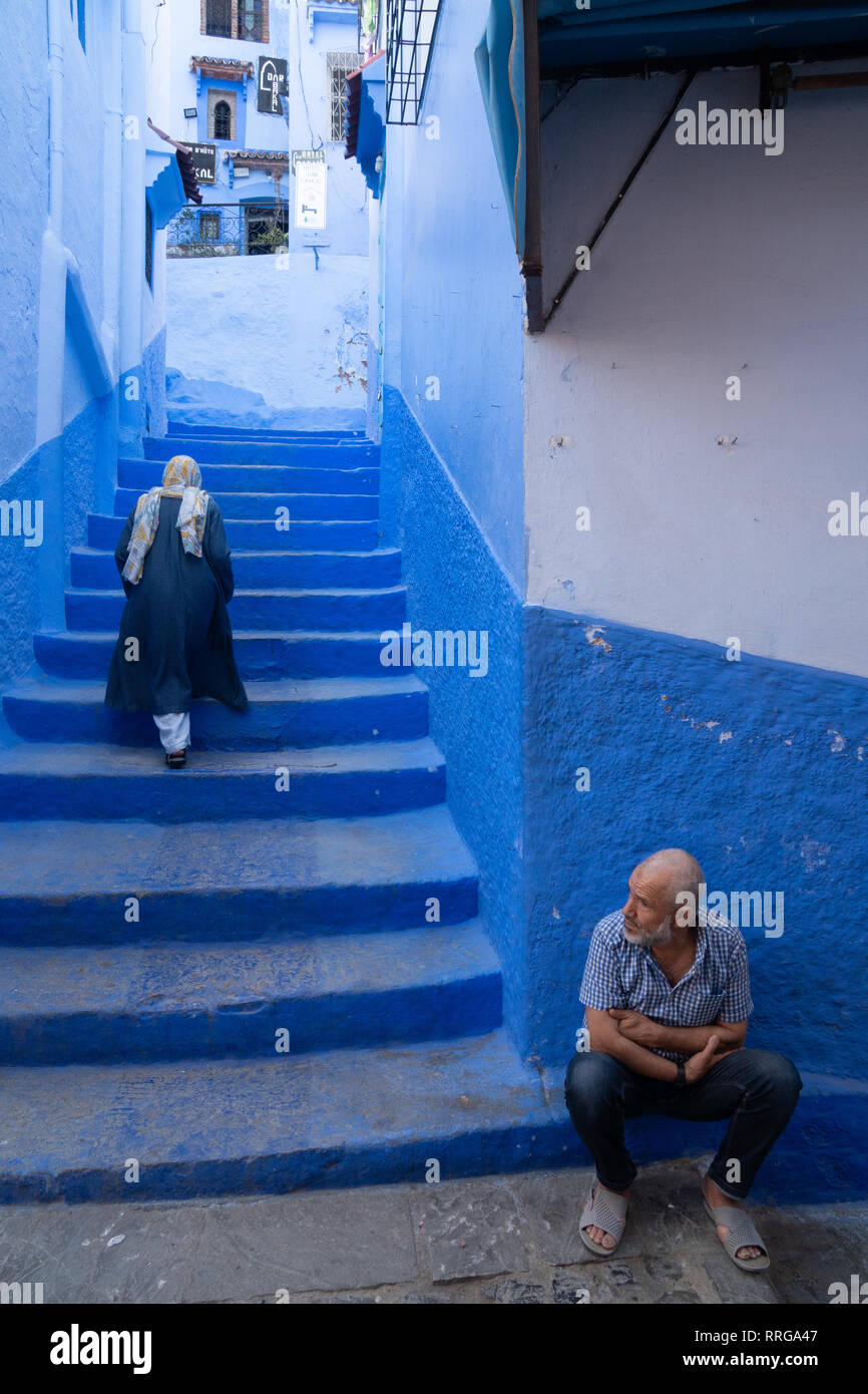 Woman in dark blue traditional clothes climbing up the stairs of alleyway, man seated in the foreground, Chefchaouen, Morocco, North Africa, Africa Stock Photo