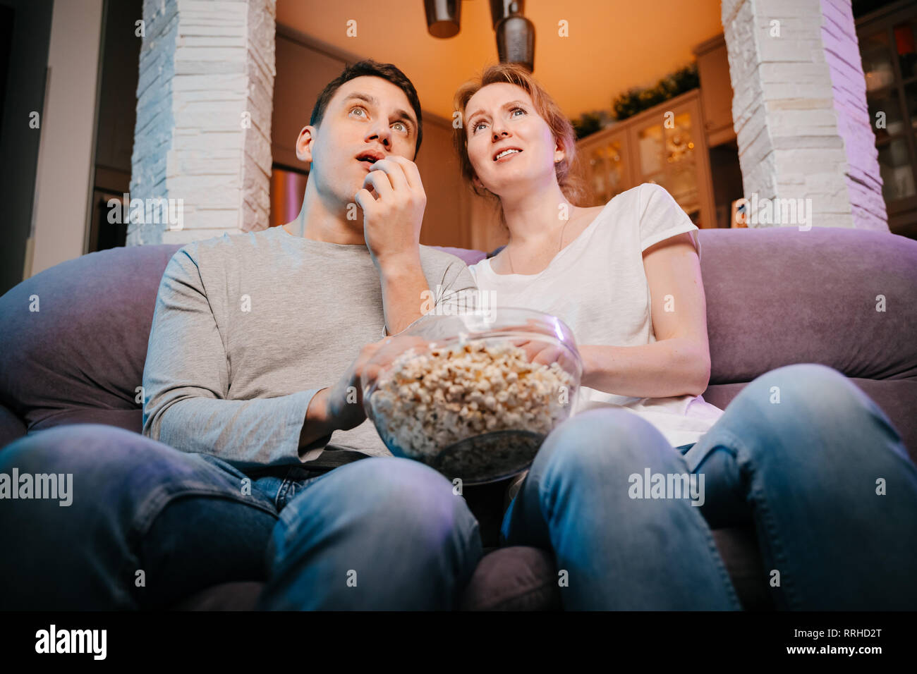 Young couple eating popcorn and watching a movie at home on the couch, scared. Stock Photo