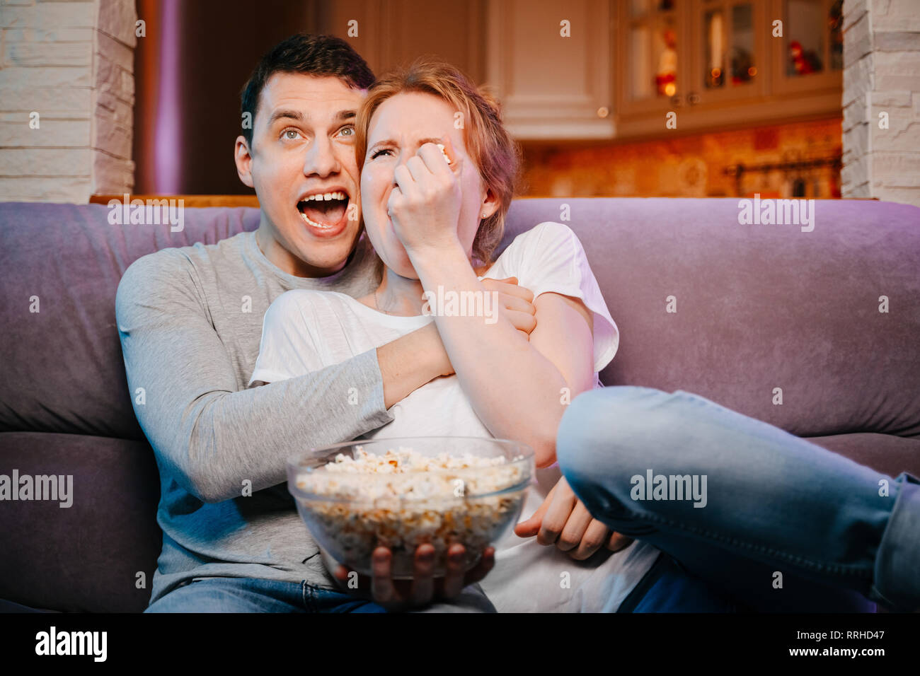 Young couple eating popcorn and watching a movie at home on the couch, scared. Stock Photo