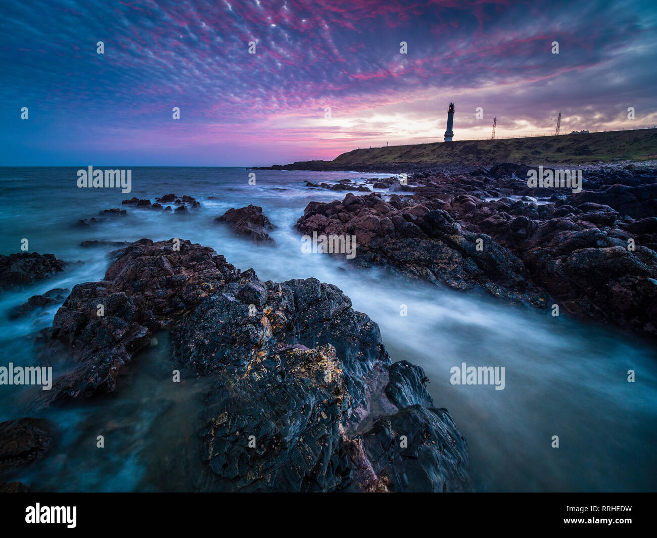 Girdle Ness lighthouse in Aberdeen during sunrise with rocks and waves in the foreground. Stock Photo