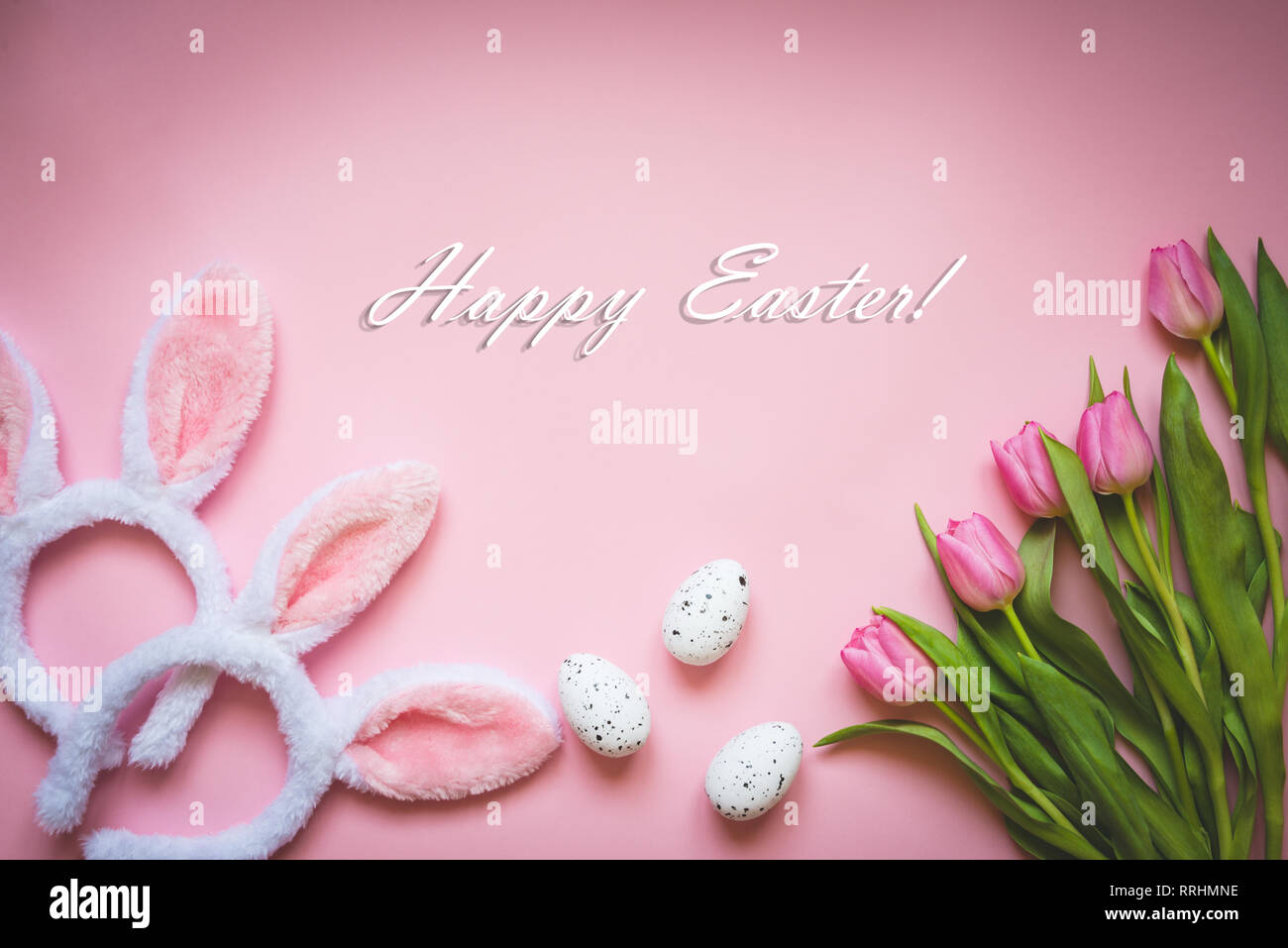 Top view of Easter eggs, pink tulips and two white fluffy bunny ears over pink background. Easter concept background. Happy Easter text. Stock Photo