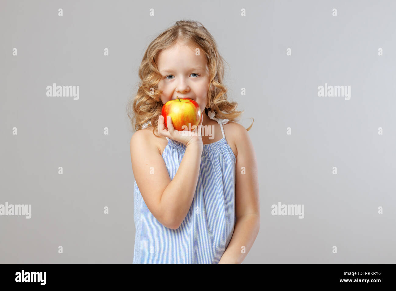 Adorable little girl child bites red apple. Gray background, studio, portrait Stock Photo