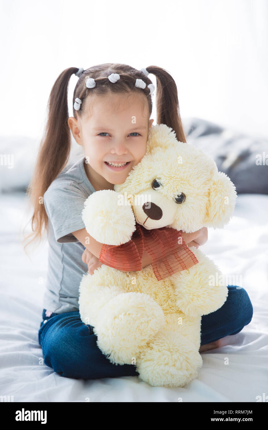 Adorable smiling littlegirl sitting on a bed hugging a white teddy bear ...