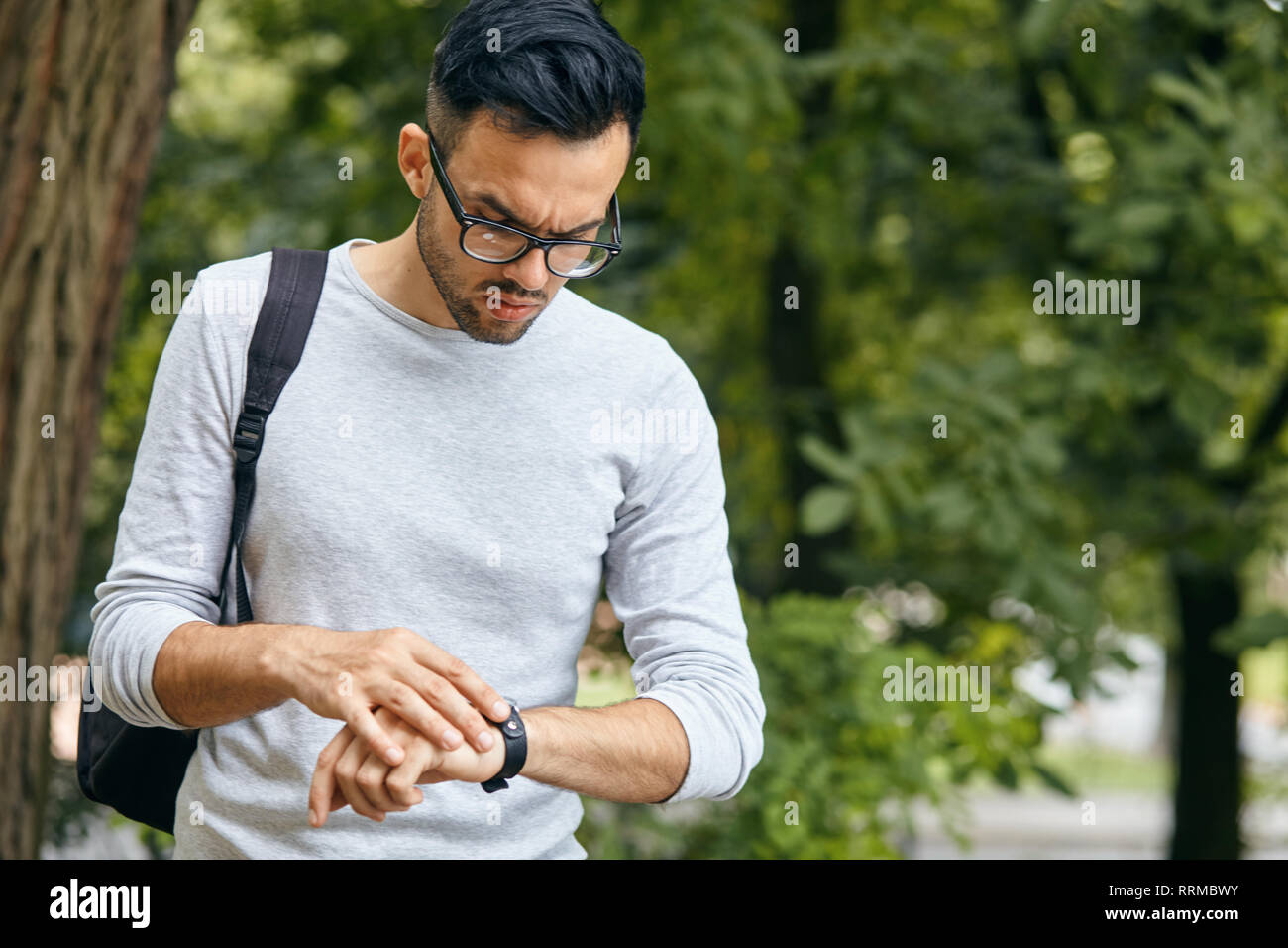 Portrait of smart responsible man with modern hairdo looking at watch on wrist over blurred park background hurry up for meeting. Management employmen Stock Photo
