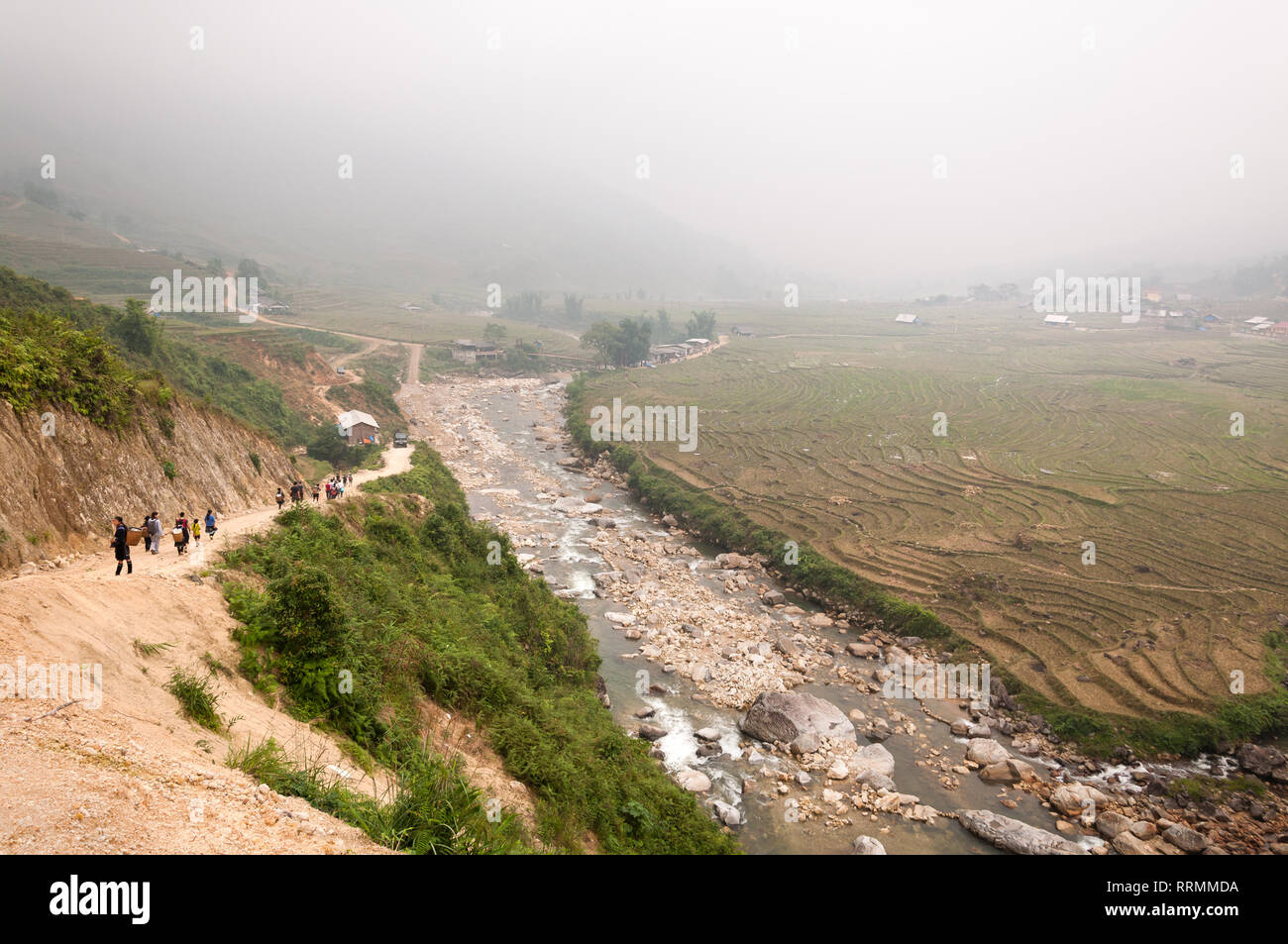 Local guides and tourists walk down a hill by a river and terraced fields on a foggy day, Sa Pa, Vietnam Stock Photo