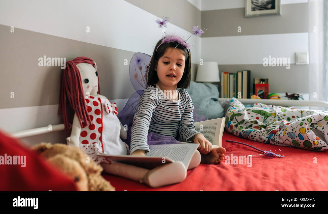Girl disguised as a butterfly teaching her doll to read Stock Photo