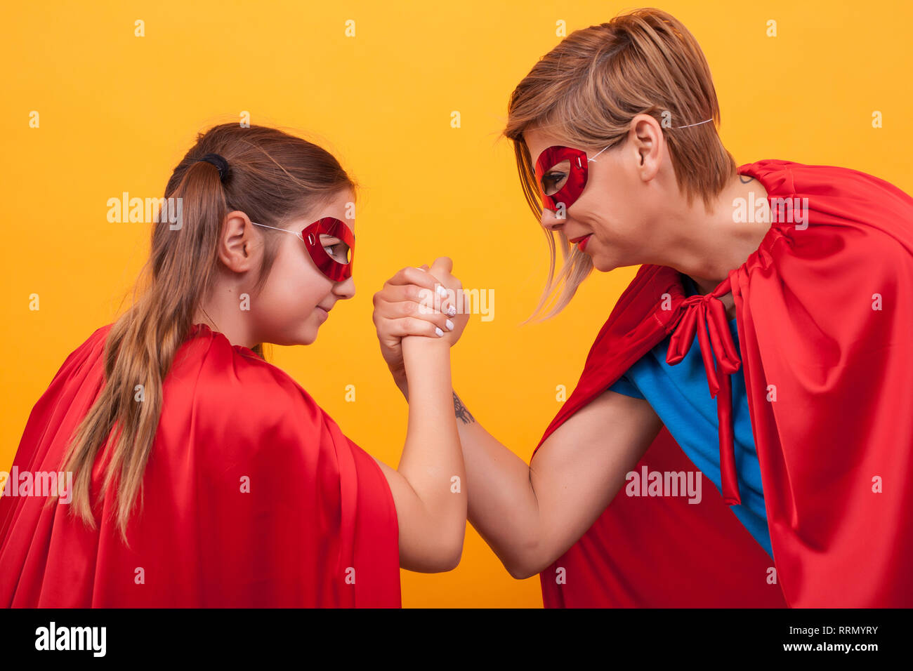 Mother and daughter dressed like superheroes playing arm wrestling over yellow background. Super mom. Super daughter. Red cape. Red mask. Parenting. Stock Photo