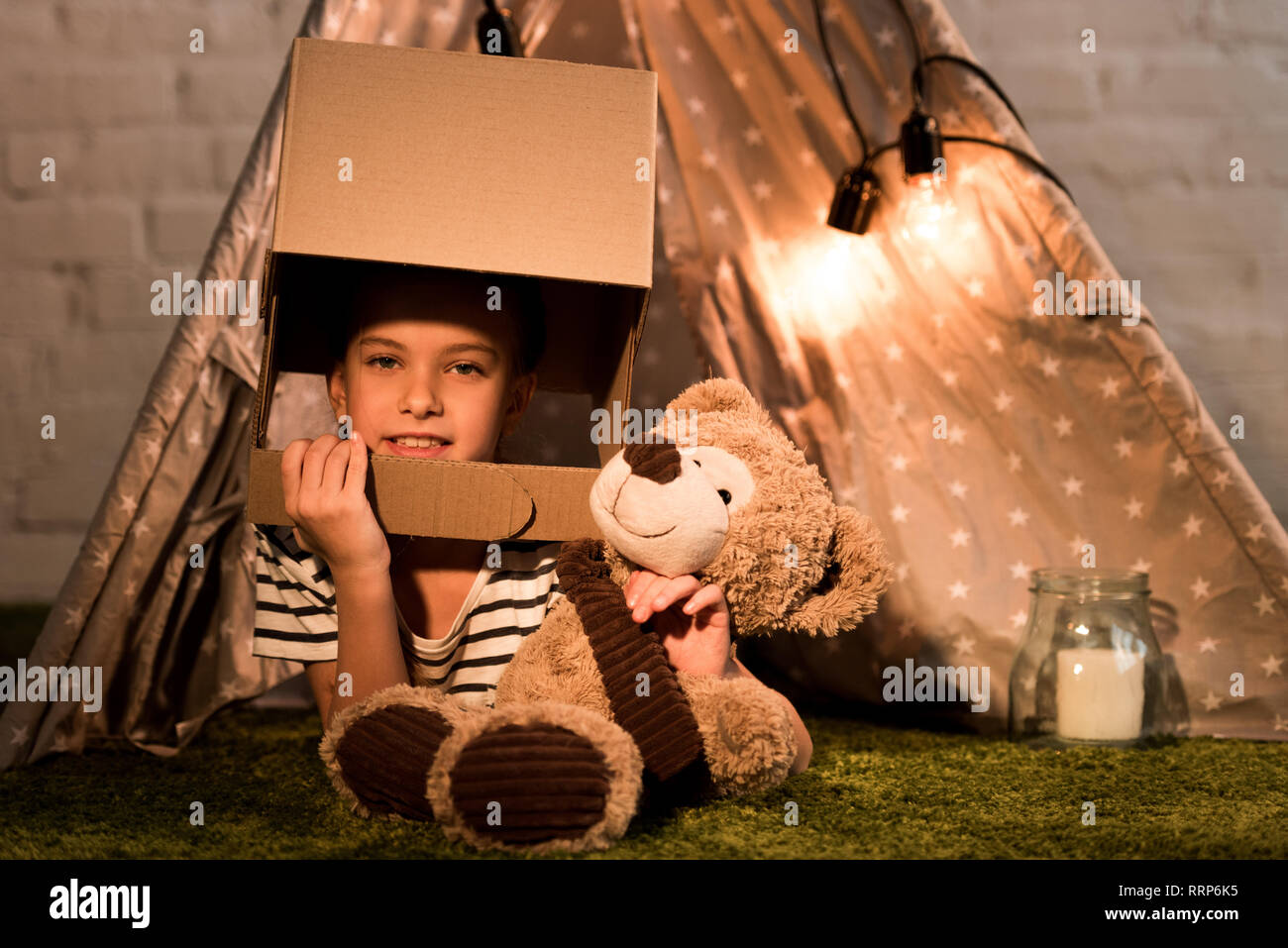 Curious kid in cardboard helmet lying on carpet with teddy bear Stock Photo