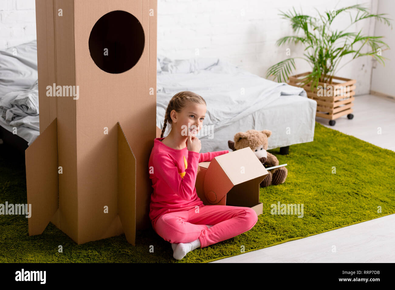 Curious kid in pink clothes sitting on carpet near cardboard rocket Stock Photo