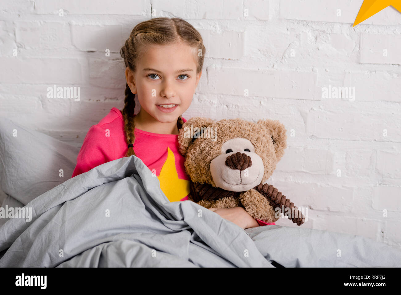 Curious kid with braids holding teddy bear in bed Stock Photo