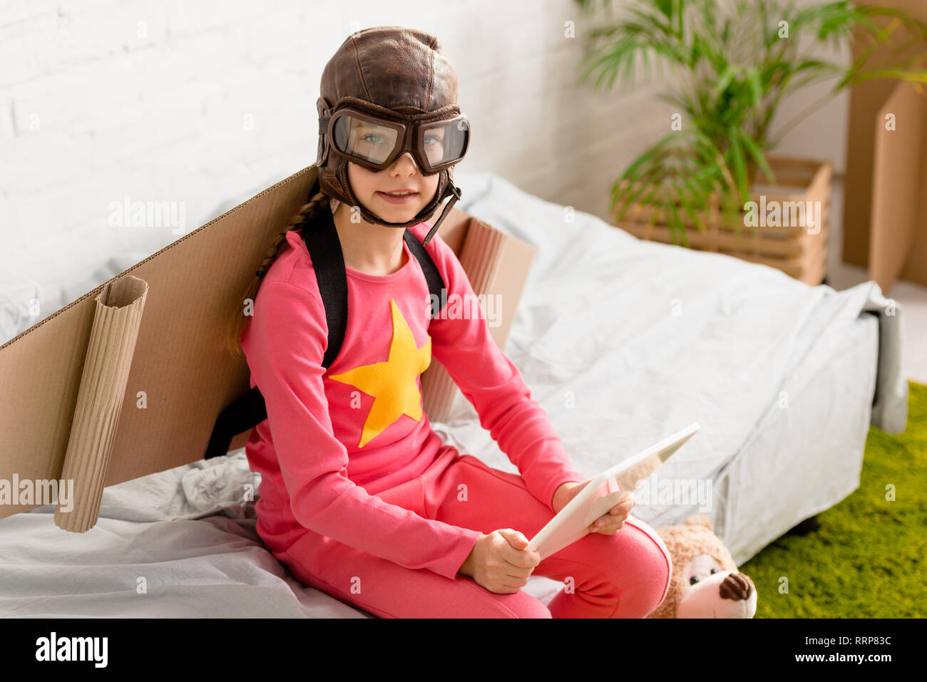 Curious kid in flight helmet sitting on bed and holding digital tablet Stock Photo
