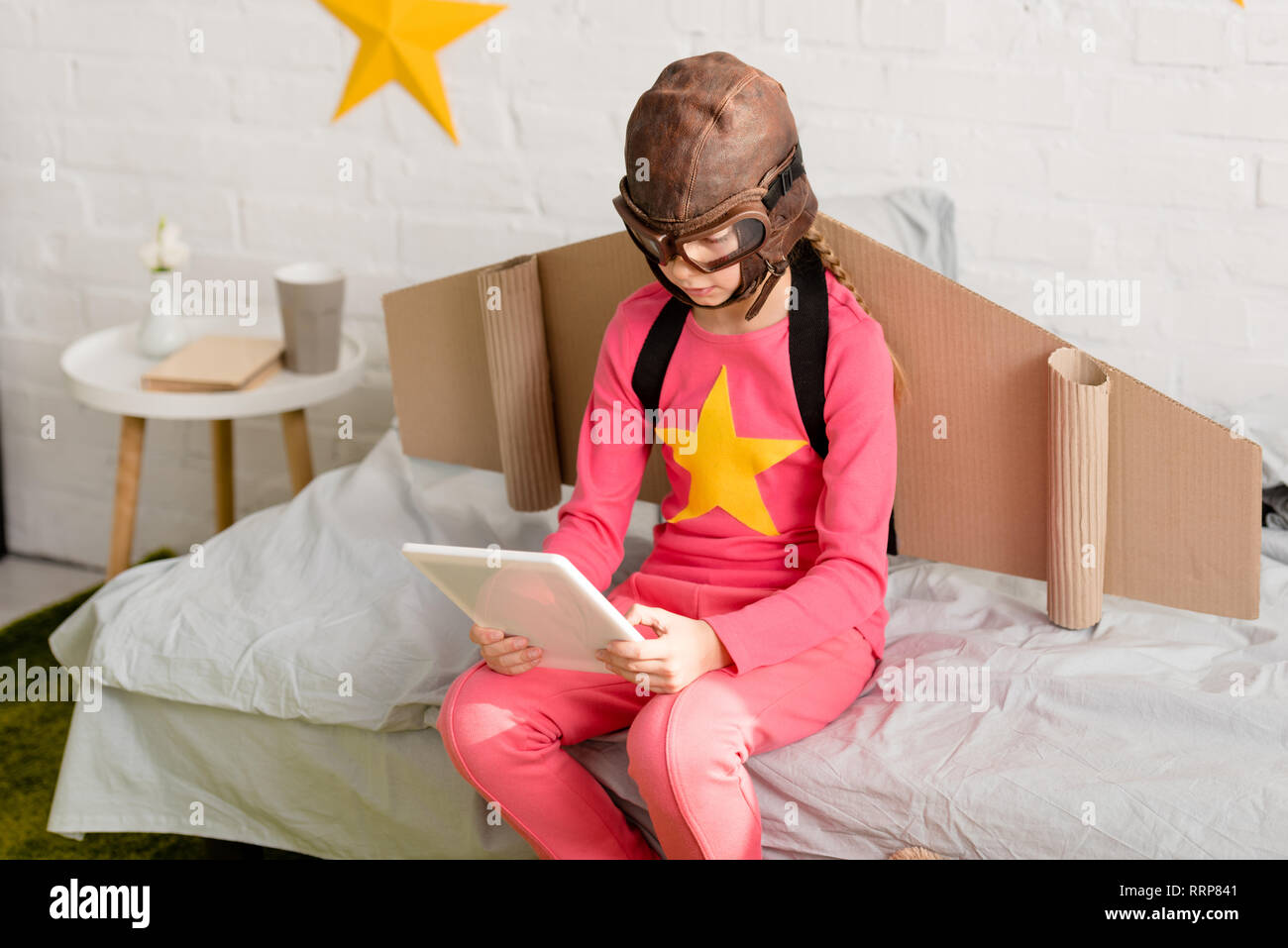 Kid in pink clothes and flight helmet using digital tablet while sitting on bed Stock Photo