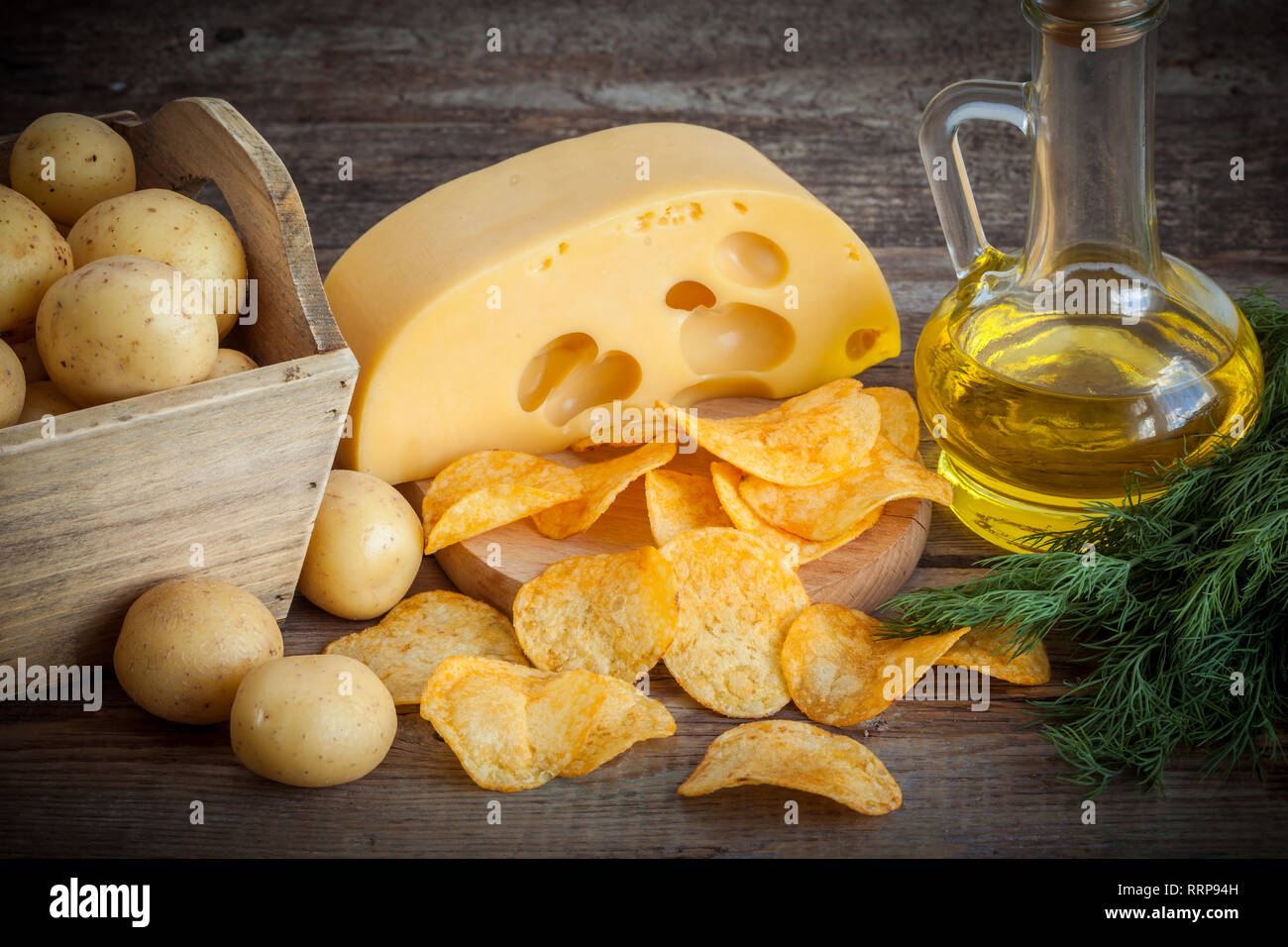 Crispy potato chips with cheese, potatoes in wooden box and oil for frying on kitchen table. Stock Photo