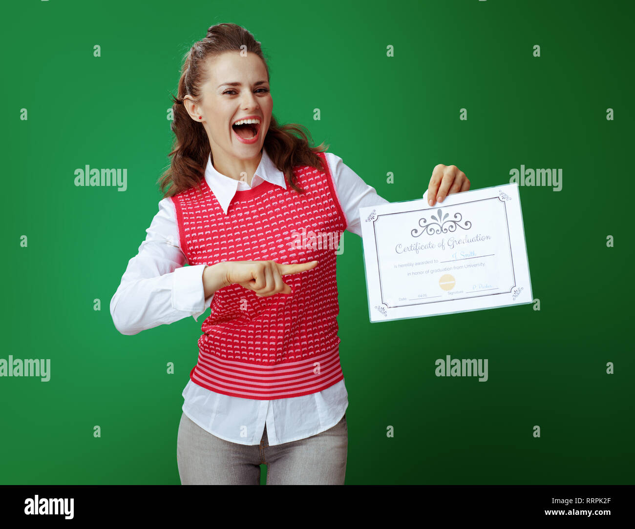 smiling modern student in grey jeans and pink sleeveless shirt pointing at Certificate of Graduation against chalkboard green background. Stock Photo