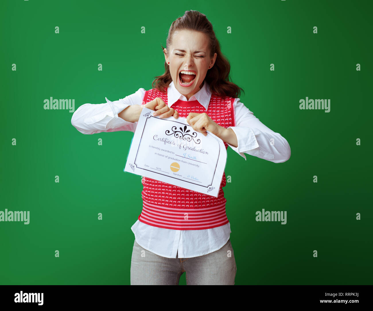 annoyed fit student woman in grey jeans and pink sleeveless shirt tearing Certificate of Graduation isolated on chalkboard green. Useless degree. Educ Stock Photo