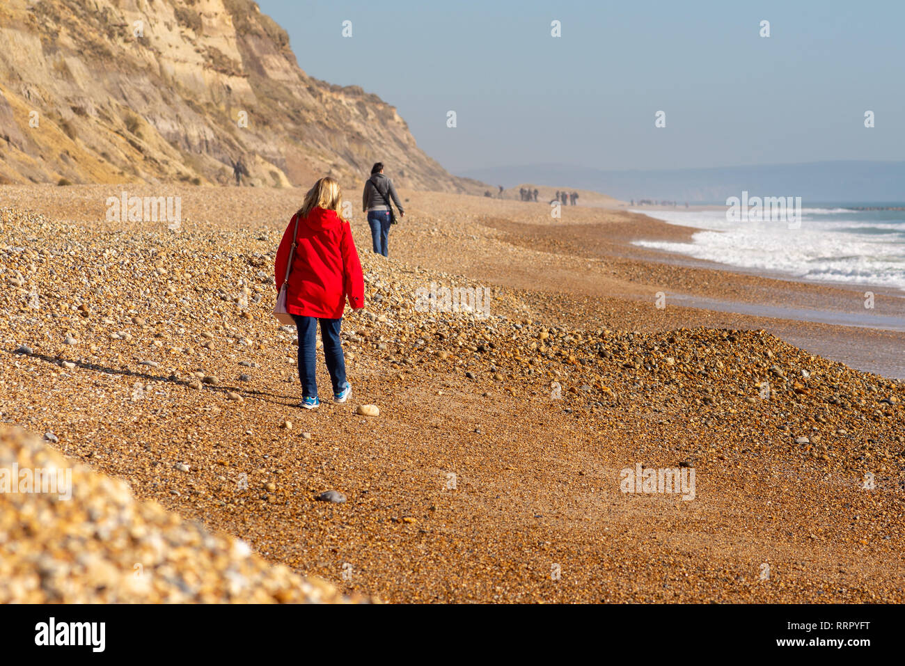 People walking along the pebbly beach at Hengistbury Head, Christchurch, Dorset, England, UK, warm February sunshine, 2019. Woman in a red coat. Stock Photo