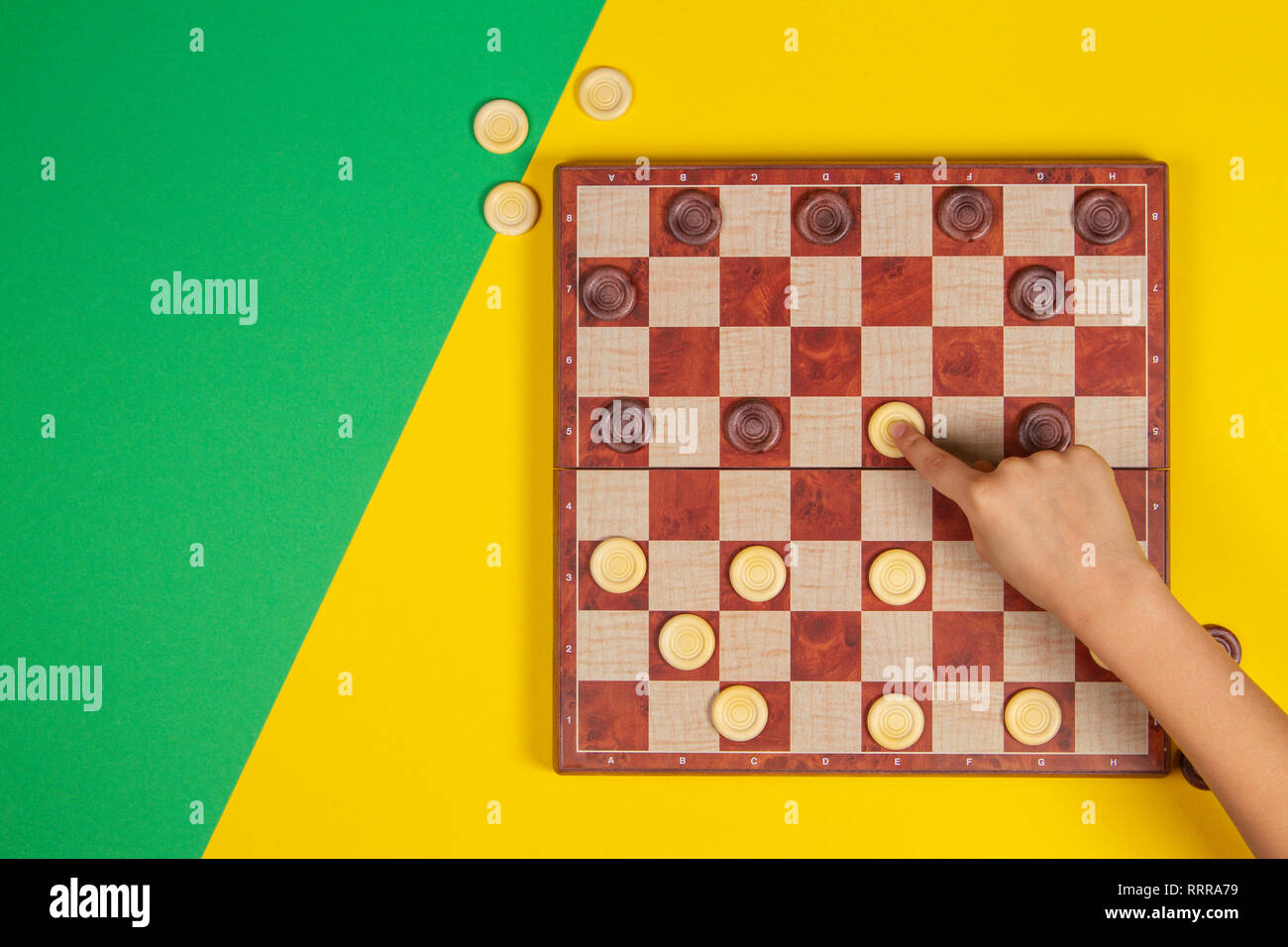 Child hand playing checkers on checker board game over yellow and green background, top view Stock Photo