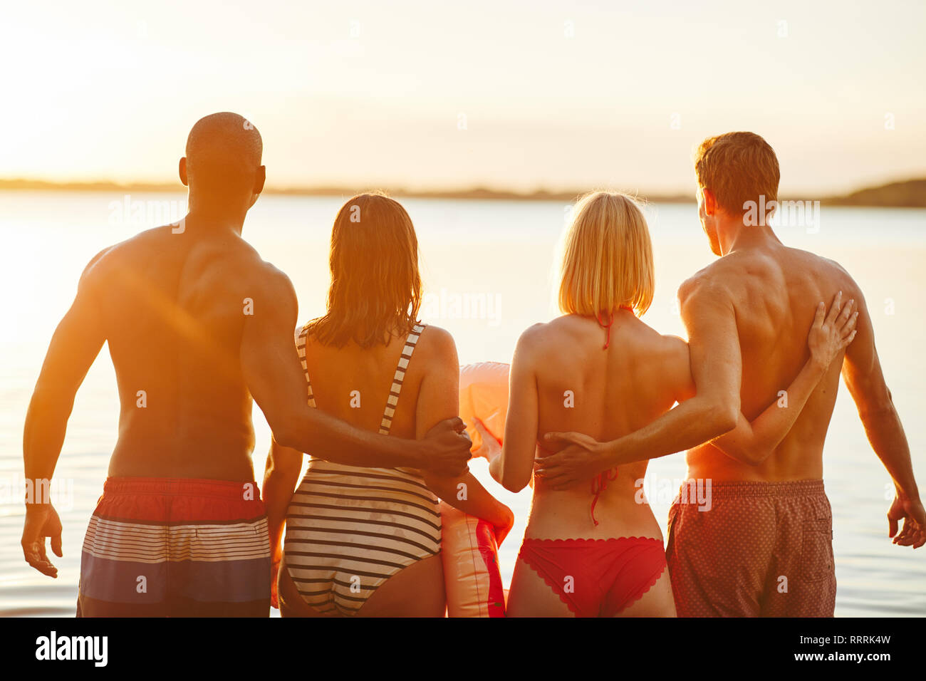 Rearview of two diverse young couples in bathing suits standing arm in arm together in a lake watching the sunset Stock Photo