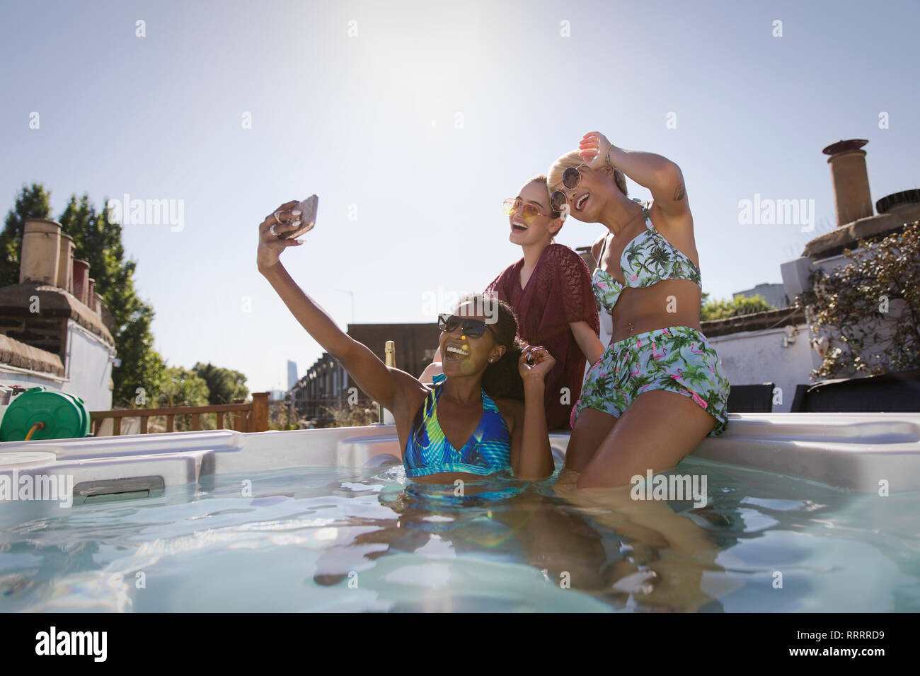 Happy, carefree young women friends in bikinis taking selfie with camera phone in sunny, rooftop hot tub Stock Photo