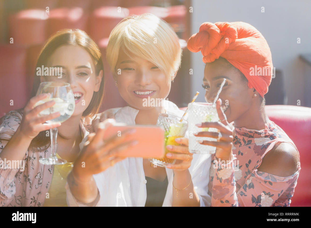 Young women friends drinking cocktails and taking selfie with smart phone in sunny bar Stock Photo