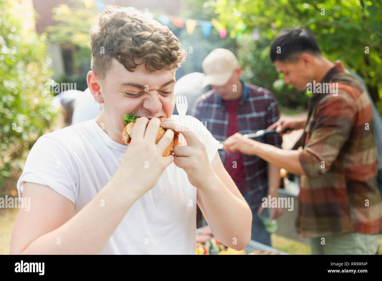 Hungry teenage boy eating hamburger at backyard barbecue Stock Photo