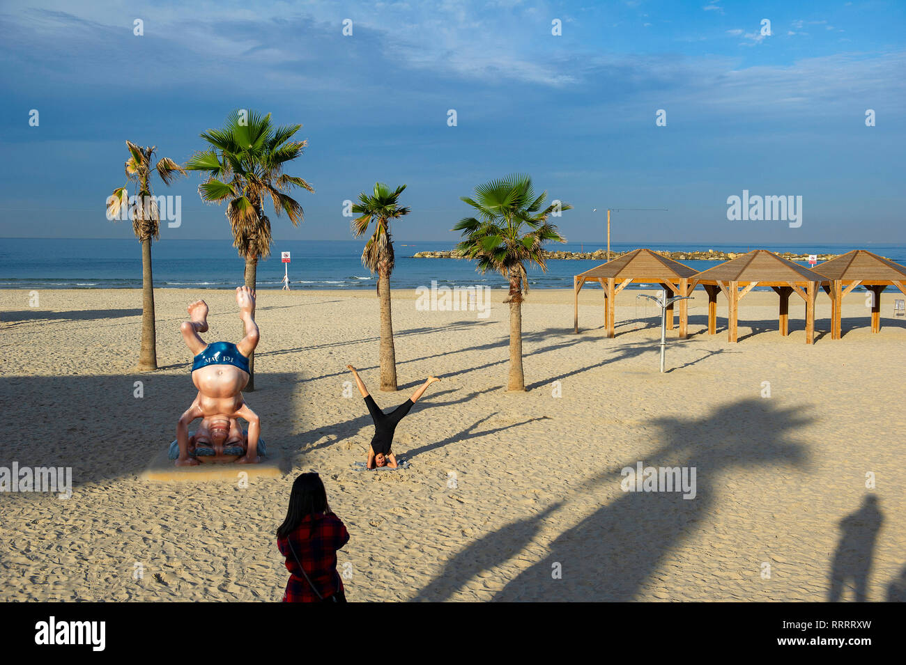 Young woman taking a picture of her friend doing yoga next to the Ben Gurion statue on the beach in Tel Aviv, Israel Stock Photo