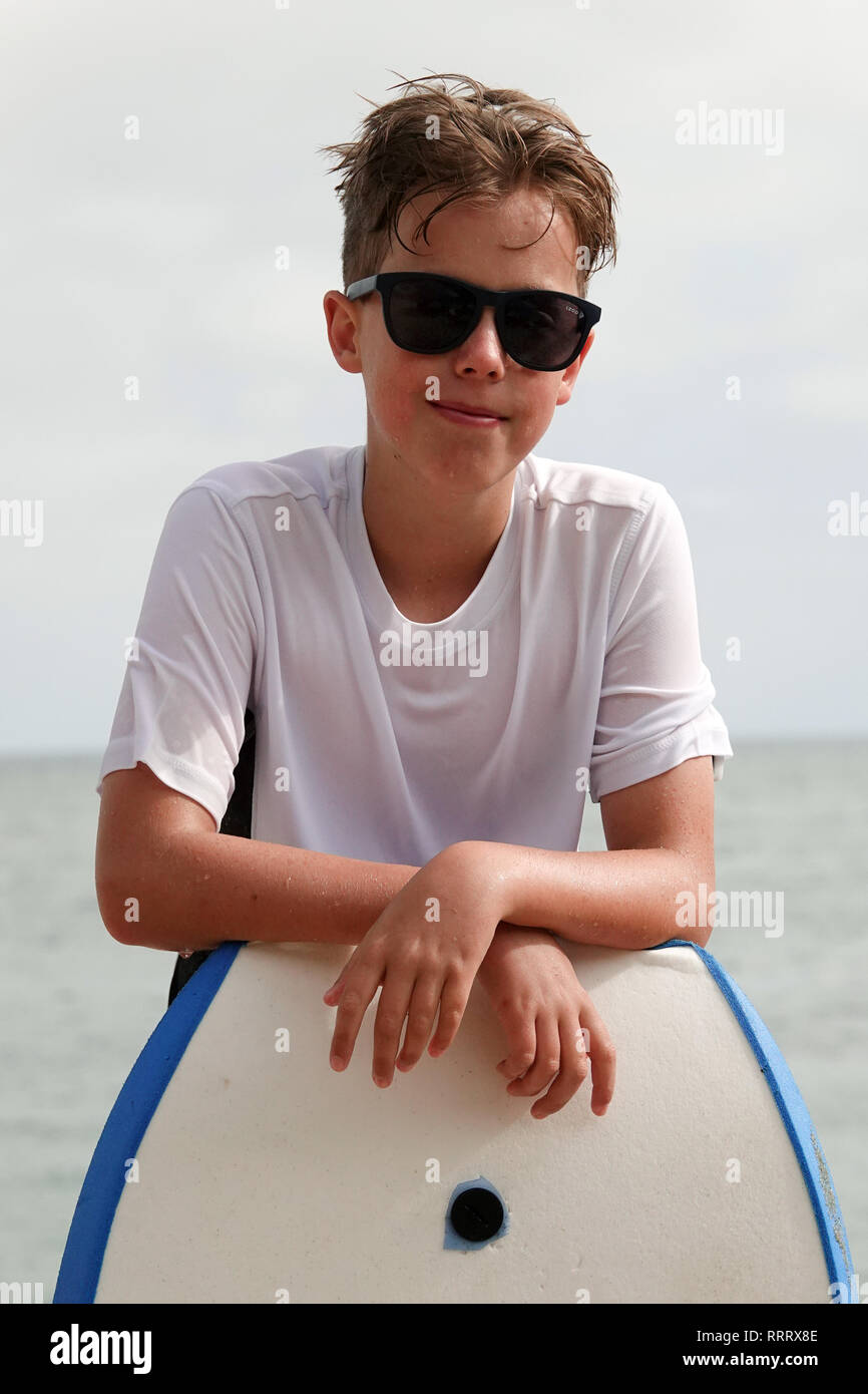A young surfer boy poses for a portrait after hitting the waves while wearing sunglasses and leaning on a boogie board. Stock Photo