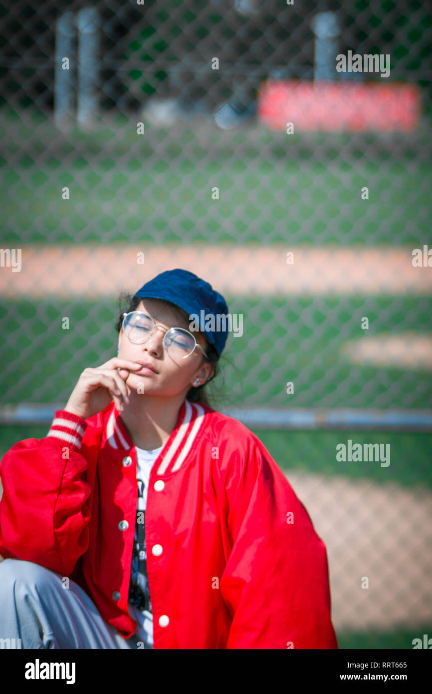 Young tennager girl model in the seat of a baseball stadium wearing red bomber jacket, hipster rounded glasses, white pants and sneakers Stock Photo