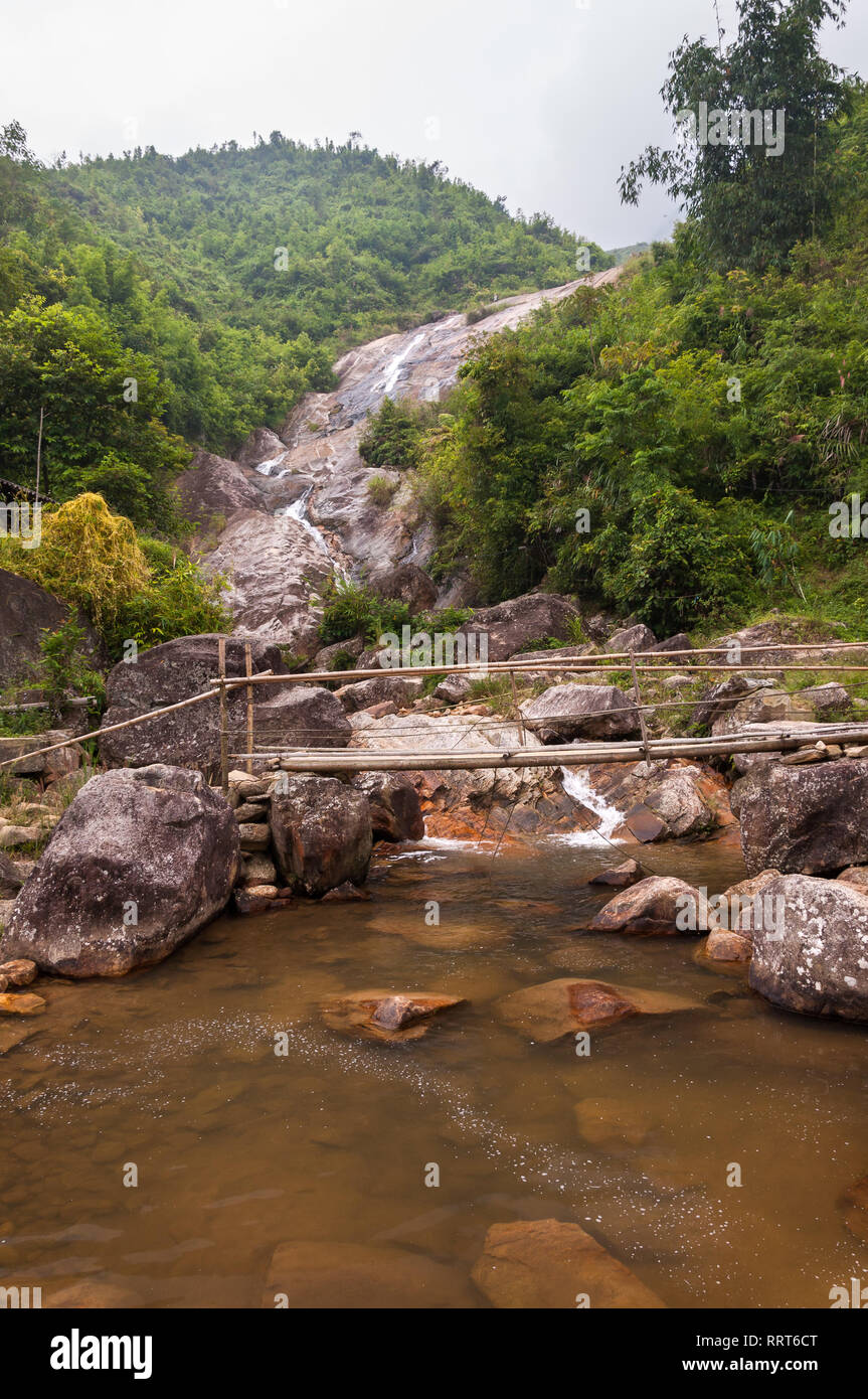 A small bamboo bridge crosses a small river in front of a waterfall, Sa Pa, Vietnam Stock Photo