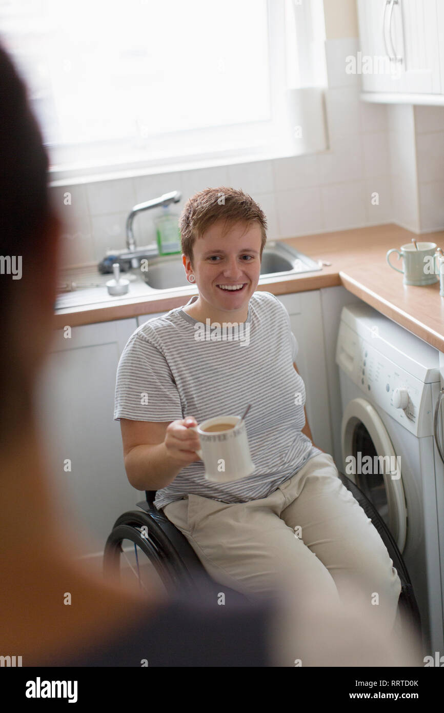 Smiling young woman in wheelchair drinking tea in apartment kitchen Stock Photo