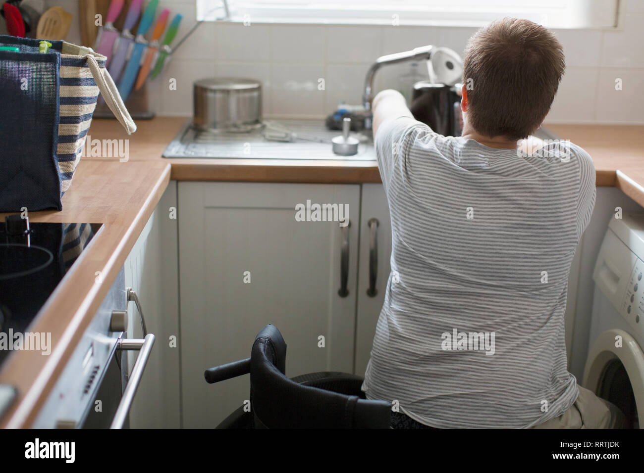 Young woman in wheelchair filling kettle for tea at apartment kitchen sink Stock Photo