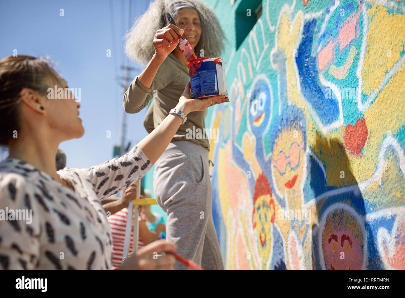 Female volunteers painting vibrant community mural on sunny urban wall Stock Photo