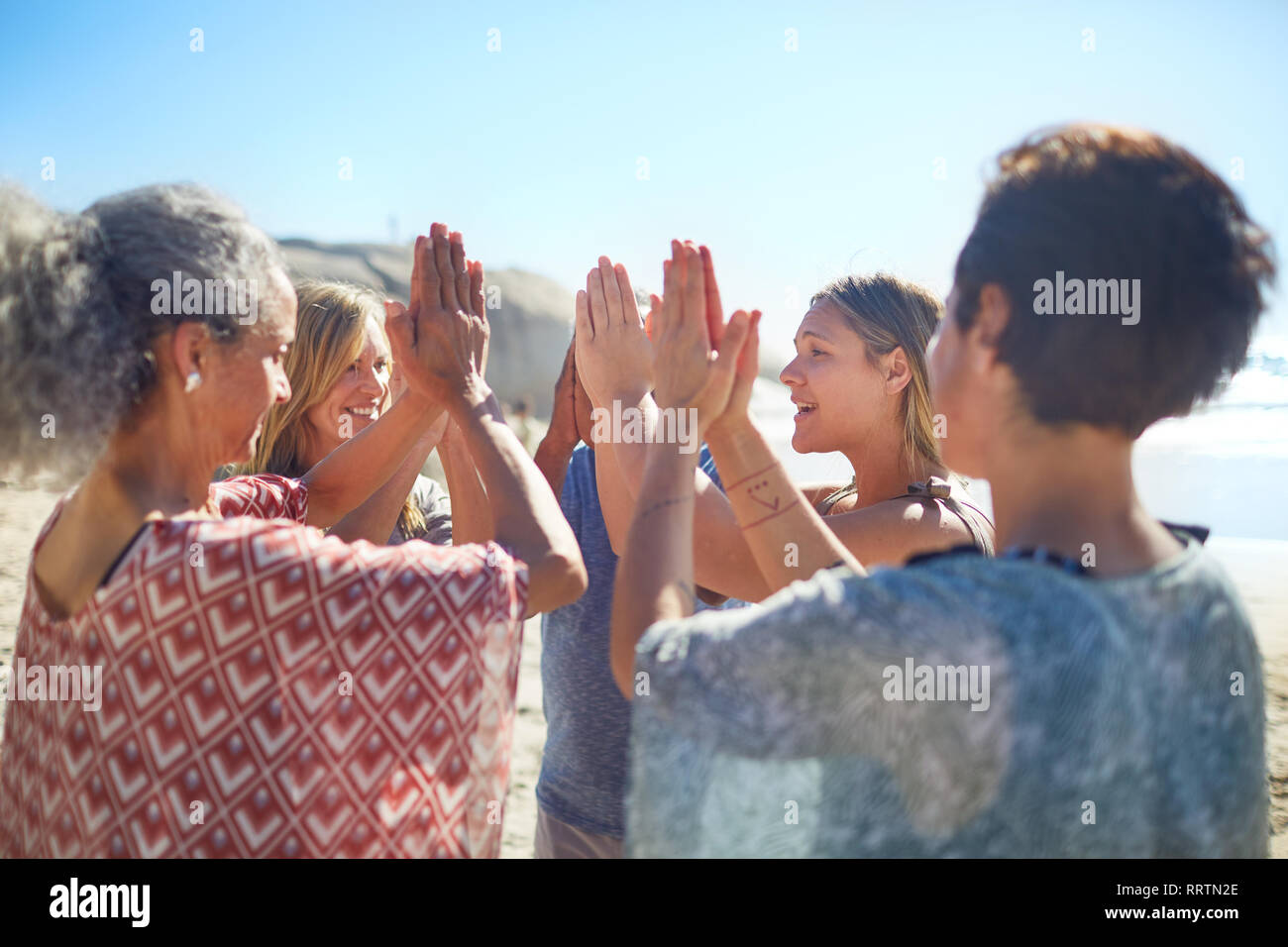 Group standing in circle with hands clasped on sunny beach during yoga retreat Stock Photo