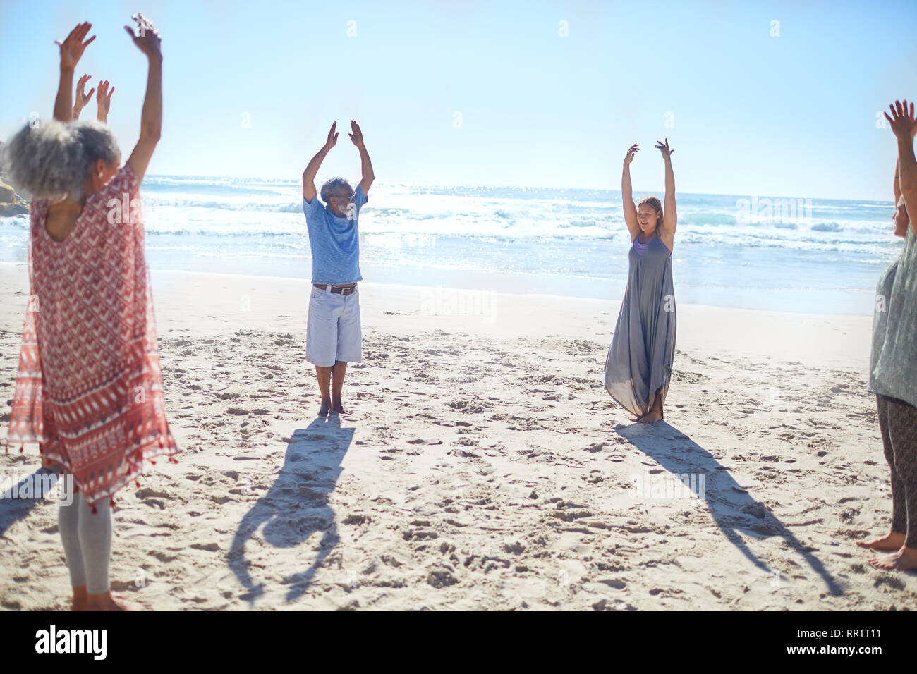 Group standing with arms raised in circle on sunny beach during yoga retreat Stock Photo