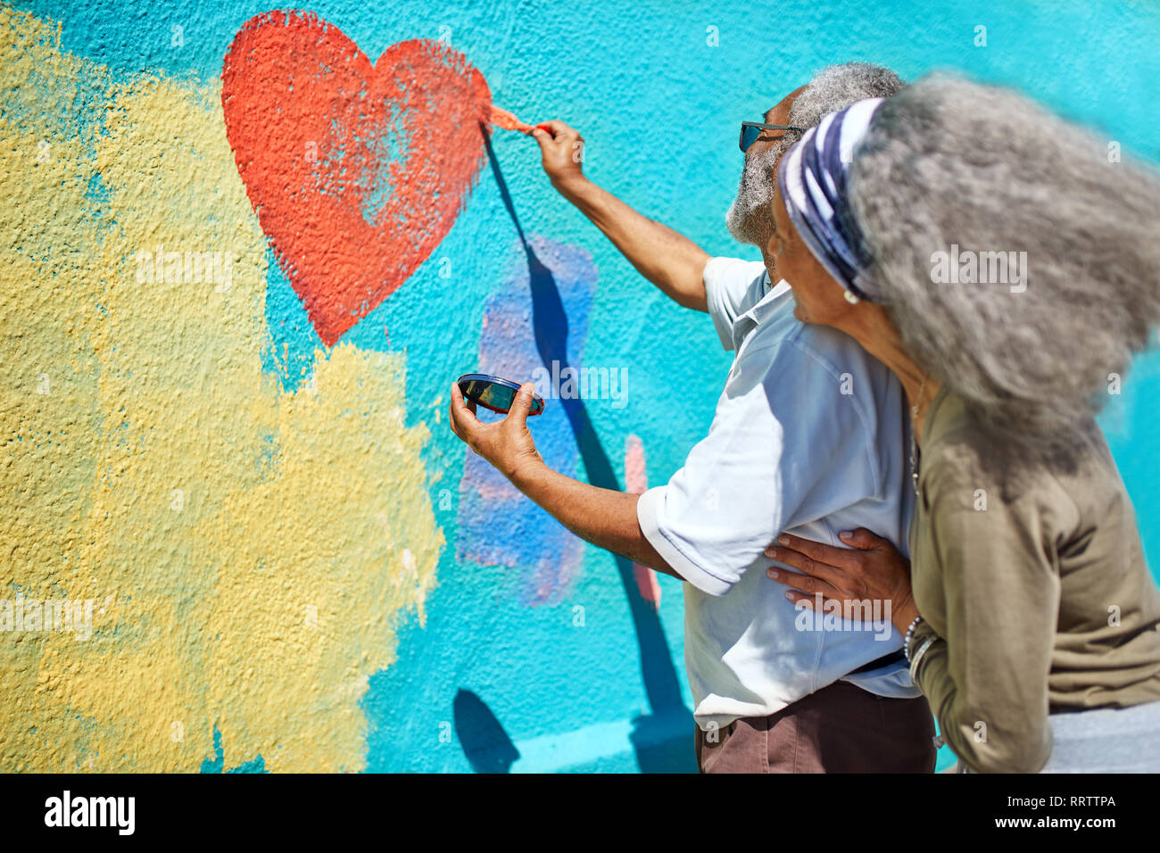 Senior couple painting heart-shape mural on sunny wall Stock Photo