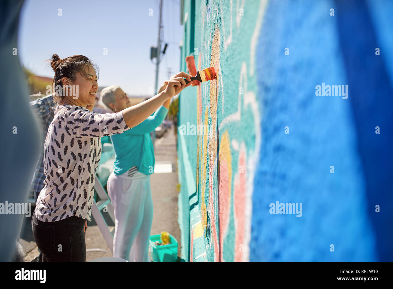 Women painting vibrant mural on sunny urban wall Stock Photo